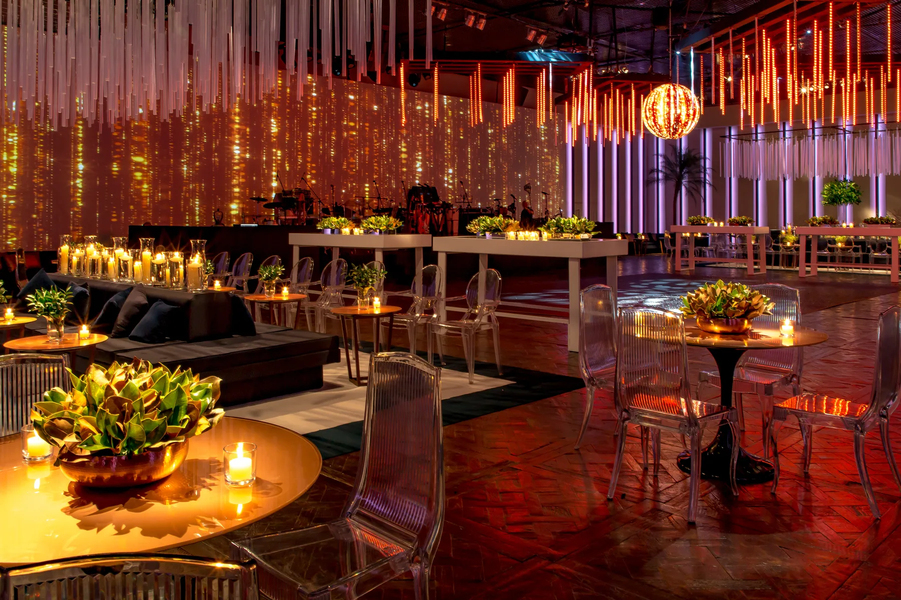 Gala ballroom in warm amber light with hanging acrylic rods, candlelit lounge seating, clear acrylic chairs and a band stage
