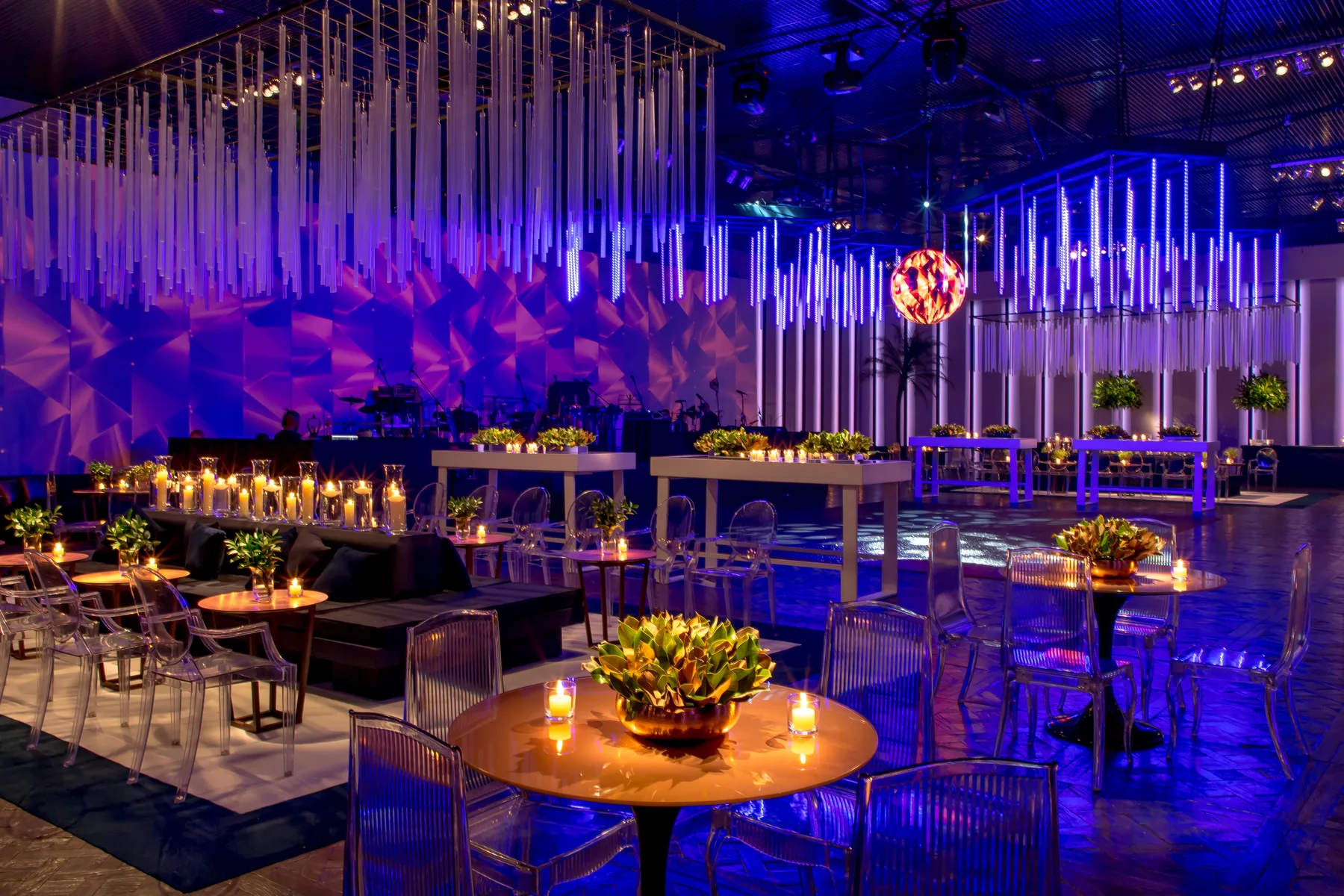 The same ballroom in blue and violet light, with hanging acrylic rods, a faceted wall, lounge seating and a band stage