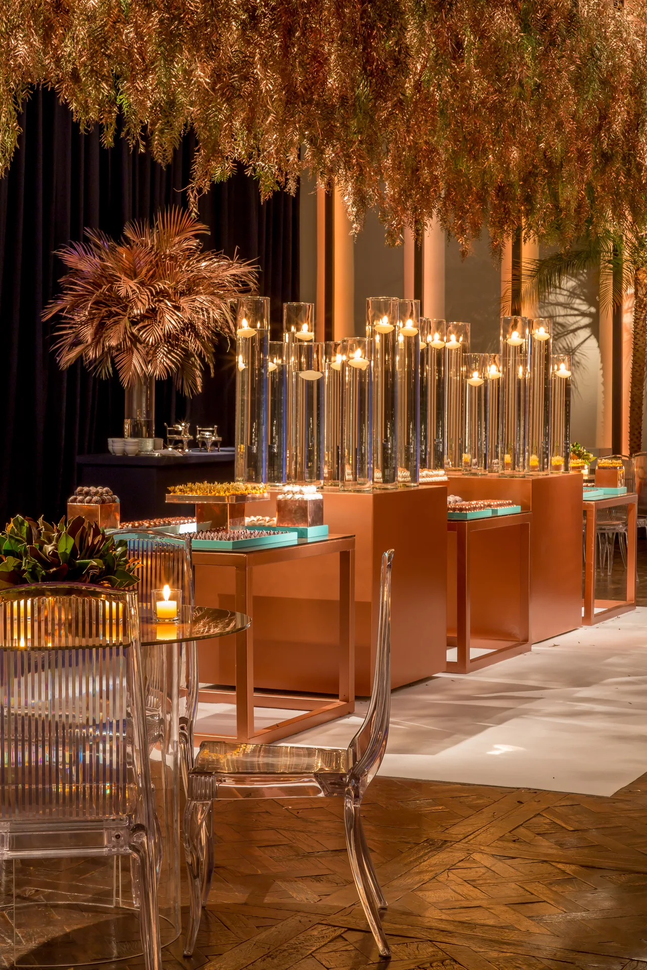 Copper dessert tables lit by tall glass candle cylinders, with gilded foliage hanging above