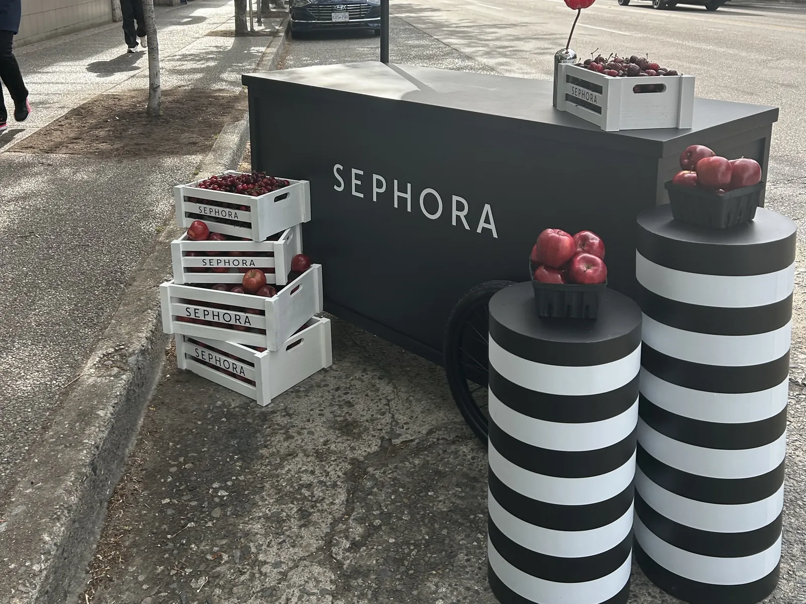 Sephora x Kits smoothie bar cart with fruit crates and black-and-white striped plinths holding apples, under a scalloped umbrella on West 4th Avenue